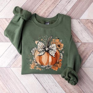 May include: An olive green sweatshirt featuring a watercolor design of a pumpkin with a polka dot bow and floral accents. The pumpkin is orange, and the flowers are shades of orange and white. The sweatshirt is folded on a wooden surface.