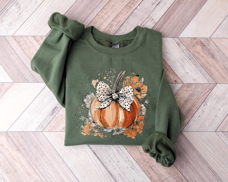 May include: An olive green sweatshirt featuring a watercolor design of a pumpkin with a polka dot bow and floral accents. The pumpkin is orange, and the flowers are shades of orange and white. The sweatshirt is folded on a wooden surface.
