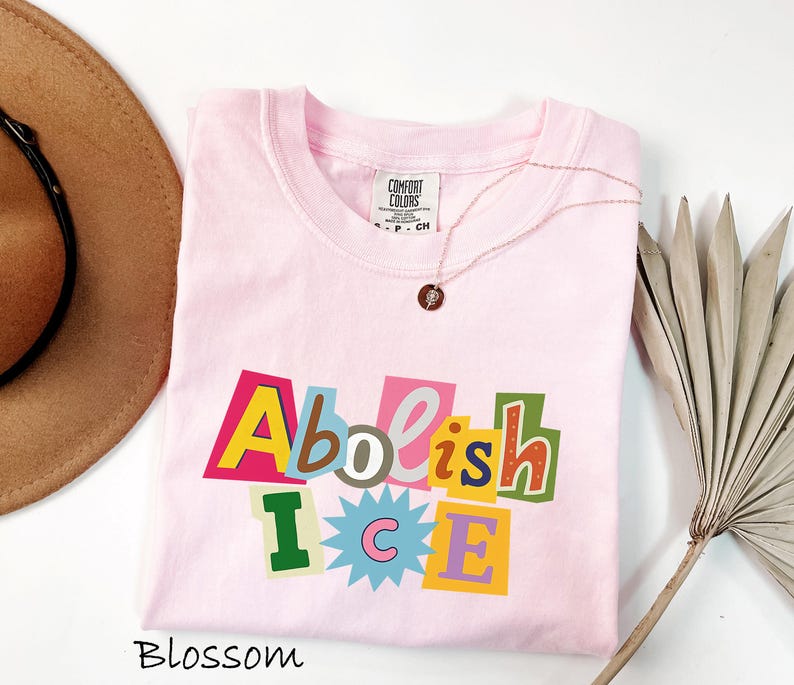May include: A light pink Comfort Colors t-shirt with the words "Abolish ICE" in a colorful, cut-out style design. A gold necklace with a small pendant rests on the shirt. A brown hat is visible in the upper left corner.