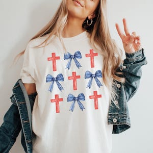 May include: White t-shirt featuring a repeated design of red crosses and blue bows.