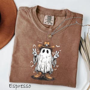 Pu&ograve; includere: T-shirt color espresso con un fantasma che indossa un cappello da cowboy e stivali, che fa il segno della pace. La grafica include una stella sul cappello e pipistrelli. Una collana d'argento con un ciondolo &egrave; drappeggiata sulla maglietta.
