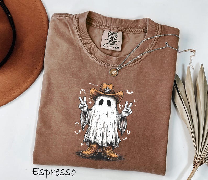 Pu&ograve; includere: T-shirt color espresso con un fantasma che indossa un cappello da cowboy e stivali, che fa il segno della pace. La grafica include una stella sul cappello e pipistrelli. Una collana d'argento con un ciondolo &egrave; drappeggiata sulla maglietta.