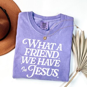 K&ouml;nnte beinhalten: Ein lavendelfarbenes T-Shirt mit dem Aufdruck "What a Friend We Have in Jesus" in Wei&szlig;. Das Shirt hat einen Used-Look und ist aus weichem, bequemem Stoff.