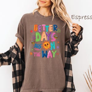 May include: Espresso-colored t-shirt with the text "BETTER DAYS ARE ON THE WAY" in colorful, playful lettering. The design includes a smiling sun, bees, and flowers. The shirt is a relaxed fit.