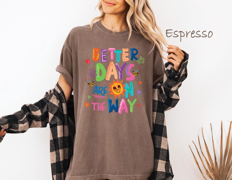 May include: Espresso-colored t-shirt with the text "BETTER DAYS ARE ON THE WAY" in colorful, playful lettering. The design includes a smiling sun, bees, and flowers. The shirt is a relaxed fit.