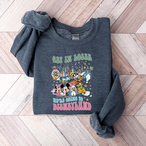 May include: A dark gray sweatshirt with a colorful graphic featuring Disney characters and the text "GET IN LOSER WE'RE GOING TO DISNEYLAND" in green and pink. The design includes Mickey Mouse, Minnie Mouse, Donald Duck, and other characters.