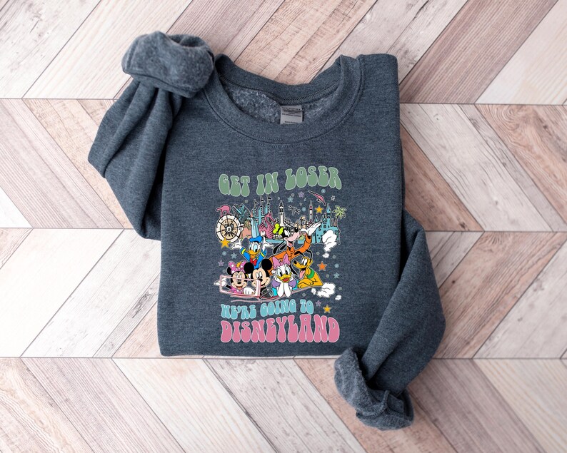 May include: A dark gray sweatshirt with a colorful graphic featuring Disney characters and the text "GET IN LOSER WE'RE GOING TO DISNEYLAND" in green and pink. The design includes Mickey Mouse, Minnie Mouse, Donald Duck, and other characters.