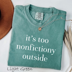 May include: A light green t-shirt with the phrase "it's too nonfictiony outside" in white. A gold necklace with a pendant rests on the shirt. The shirt is folded, with a brown hat and dried plant in the background.
