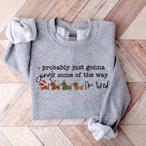May include: A heather gray sweatshirt with the text "probably just gonna jingle some of the way I'm tired." The design includes four dachshunds dressed in Christmas attire. The sweatshirt has a crew neck and long sleeves.