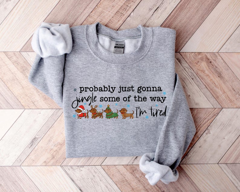 May include: A heather gray sweatshirt with the text "probably just gonna jingle some of the way I'm tired." The design includes four dachshunds dressed in Christmas attire. The sweatshirt has a crew neck and long sleeves.