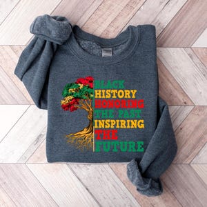 Pu&ograve; includere: Una felpa grigio scuro con la scritta "BLACK HISTORY HONORING THE PAST INSPIRING THE FUTURE" in verde, rosso e giallo. Una grafica ad albero con foglie rosse, verdi e gialle si trova sul lato sinistro del testo.