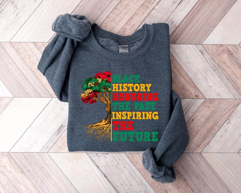 Pu&ograve; includere: Una felpa grigio scuro con la scritta "BLACK HISTORY HONORING THE PAST INSPIRING THE FUTURE" in verde, rosso e giallo. Una grafica ad albero con foglie rosse, verdi e gialle si trova sul lato sinistro del testo.