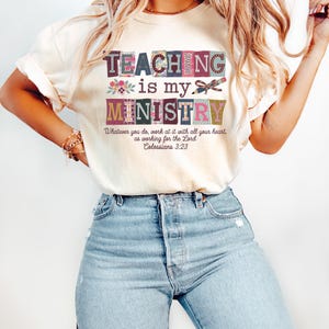 May include: Cream-colored t-shirt with the text "TEACHING is my MINISTRY" in a colorful, block-letter design. The shirt also features the Bible verse Colossians 3:23. The shirt is paired with light blue jeans.