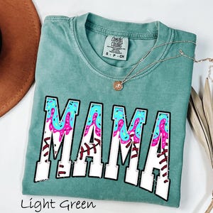May include: Light green t-shirt with the word "MAMA" in white letters with a pink, blue, and white donut glaze design. The shirt has a distressed look and is made of a soft, comfortable fabric.
