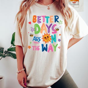 May include: Ivory-colored t-shirt with the phrase "BETTER DAYS ARE ON THE WAY" in colorful letters and whimsical illustrations. The design includes a smiling sun, bees, flowers, and hearts. The shirt is a relaxed fit.