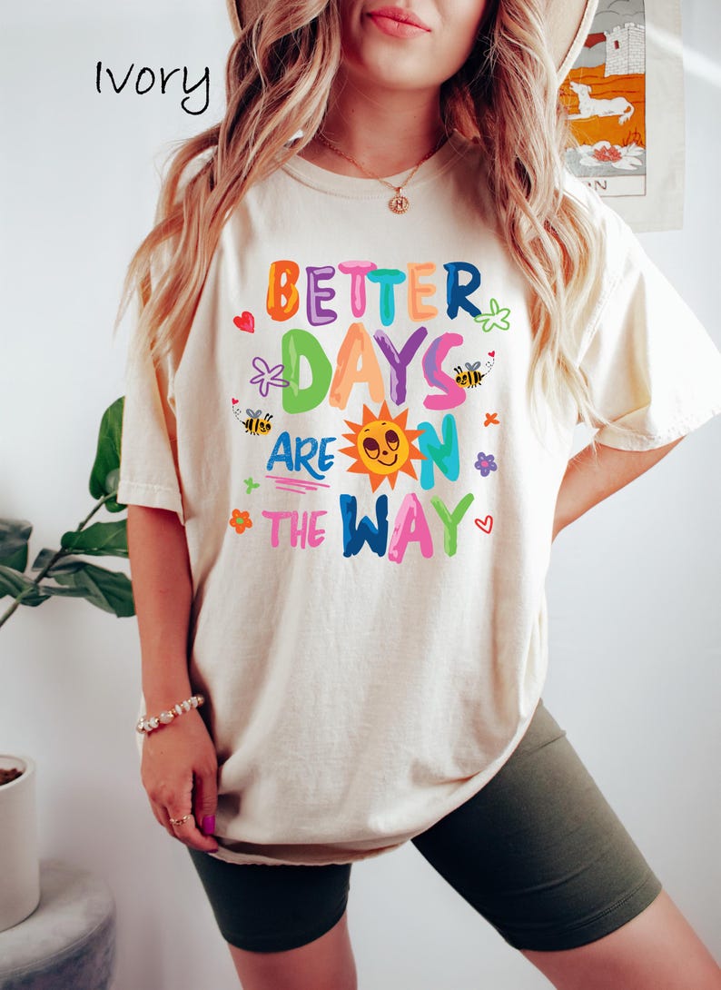 May include: Ivory-colored t-shirt with the phrase "BETTER DAYS ARE ON THE WAY" in colorful letters and whimsical illustrations. The design includes a smiling sun, bees, flowers, and hearts. The shirt is a relaxed fit.