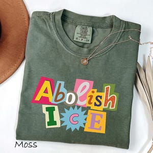 May include: Moss green t-shirt with the text "Abolish ICE" in colorful, block letter style. The shirt has a crew neck and short sleeves. A gold necklace with a small pendant is draped on the shirt.
