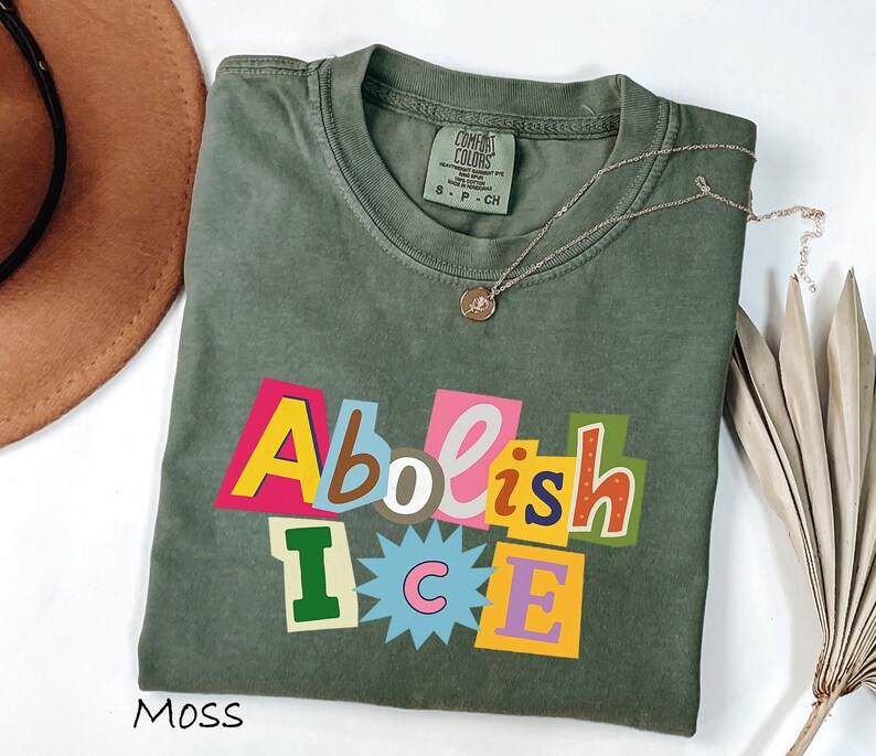 May include: Moss green t-shirt with the text "Abolish ICE" in colorful, block letter style. The shirt has a crew neck and short sleeves. A gold necklace with a small pendant is draped on the shirt.