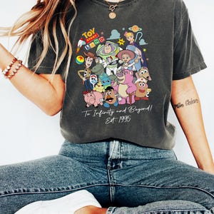 May include: Dark gray t-shirt featuring a colorful graphic of Toy Story characters. The design includes Woody, Buzz Lightyear, and other characters from the movie. Text on the shirt reads "To Infinity and Beyond! Est. 1995".