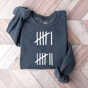 May include: A dark gray sweatshirt with white tally marks. The sweatshirt is folded on a light brown wooden surface. The tally marks are arranged in two sets, each with four vertical lines and one diagonal line.
