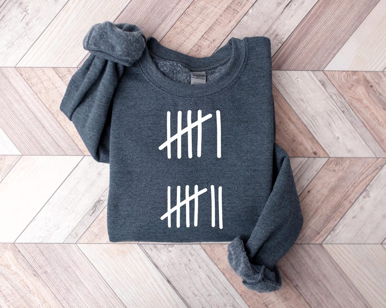 May include: A dark gray sweatshirt with white tally marks. The sweatshirt is folded on a light brown wooden surface. The tally marks are arranged in two sets, each with four vertical lines and one diagonal line.