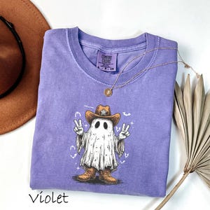 Pu&ograve; includere: T-shirt viola con un fantasma dei cartoni animati che indossa un cappello e stivali da cowboy, che fa il segno della pace. L'illustrazione del fantasma ha un'estetica vintage. La maglietta ha un girocollo e maniche corte. Una collana d'oro &egrave; drappeggiata sulla maglietta.