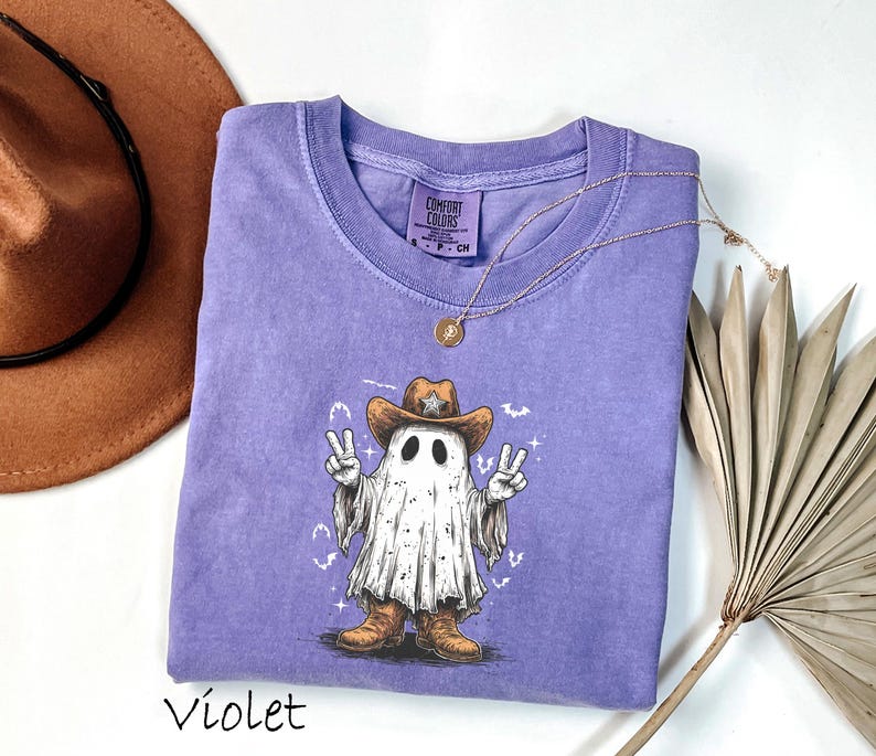 Pu&ograve; includere: T-shirt viola con un fantasma dei cartoni animati che indossa un cappello e stivali da cowboy, che fa il segno della pace. L'illustrazione del fantasma ha un'estetica vintage. La maglietta ha un girocollo e maniche corte. Una collana d'oro &egrave; drappeggiata sulla maglietta.