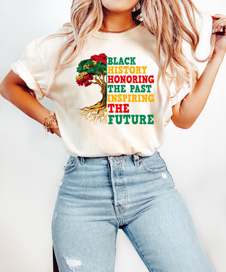Pu&ograve; includere: T-shirt color crema con grafica di un albero colorato e la scritta "BLACK HISTORY HONORING THE PAST INSPIRING THE FUTURE" in nero, rosso, giallo e verde. La maglietta &egrave; abbinata a jeans di denim blu chiaro.