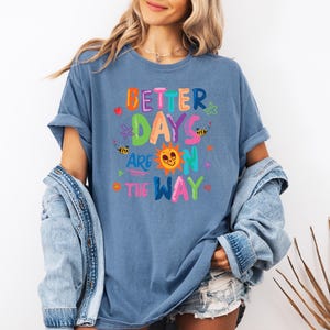 May include: A blue t-shirt with the phrase "Better Days Are On The Way" in colorful, playful lettering. The design includes a smiling sun, bees, and floral accents. The shirt is a casual, comfortable style.
