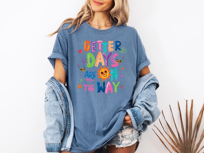 May include: A blue t-shirt with the phrase "Better Days Are On The Way" in colorful, playful lettering. The design includes a smiling sun, bees, and floral accents. The shirt is a casual, comfortable style.