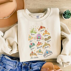 May include: Cream-colored sweatshirt featuring a colorful illustration of tiaras, each labeled with a princess's name. The design includes tiaras for Tiana, Elsa, Ariel, Rapunzel, Jasmine, Snow White, Belle, Aurora, Moana, Mulan, and Cinderella.