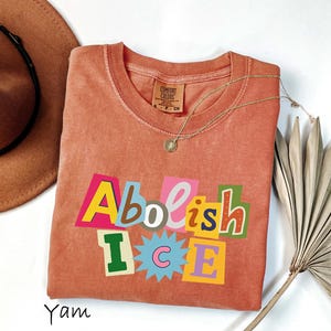 May include: A coral-colored t-shirt with the words "Abolish ICE" in a colorful, cut-out style design. The shirt is styled with a gold necklace and a brown hat.