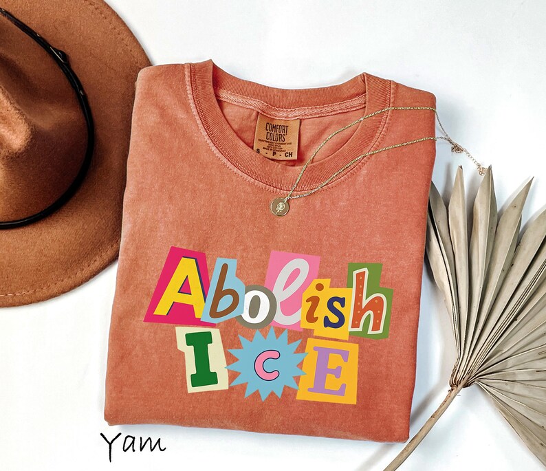 May include: A coral-colored t-shirt with the words "Abolish ICE" in a colorful, cut-out style design. The shirt is styled with a gold necklace and a brown hat.