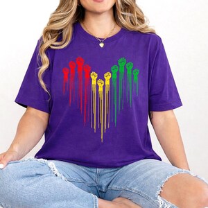 May include: A purple t-shirt featuring a heart-shaped design of raised fists in red, yellow, and green. The fists are arranged in vertical lines, creating a striking visual on the shirt.