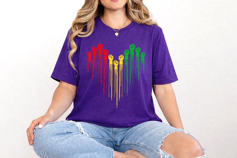 May include: A purple t-shirt featuring a heart-shaped design of raised fists in red, yellow, and green. The fists are arranged in vertical lines, creating a striking visual on the shirt.