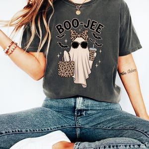 May include: Dark grey t-shirt with a Halloween graphic. The design shows a ghost wearing sunglasses and a leopard print bow, holding a leopard print bag and a coffee cup. The text "BOO-JEE" is above the ghost.