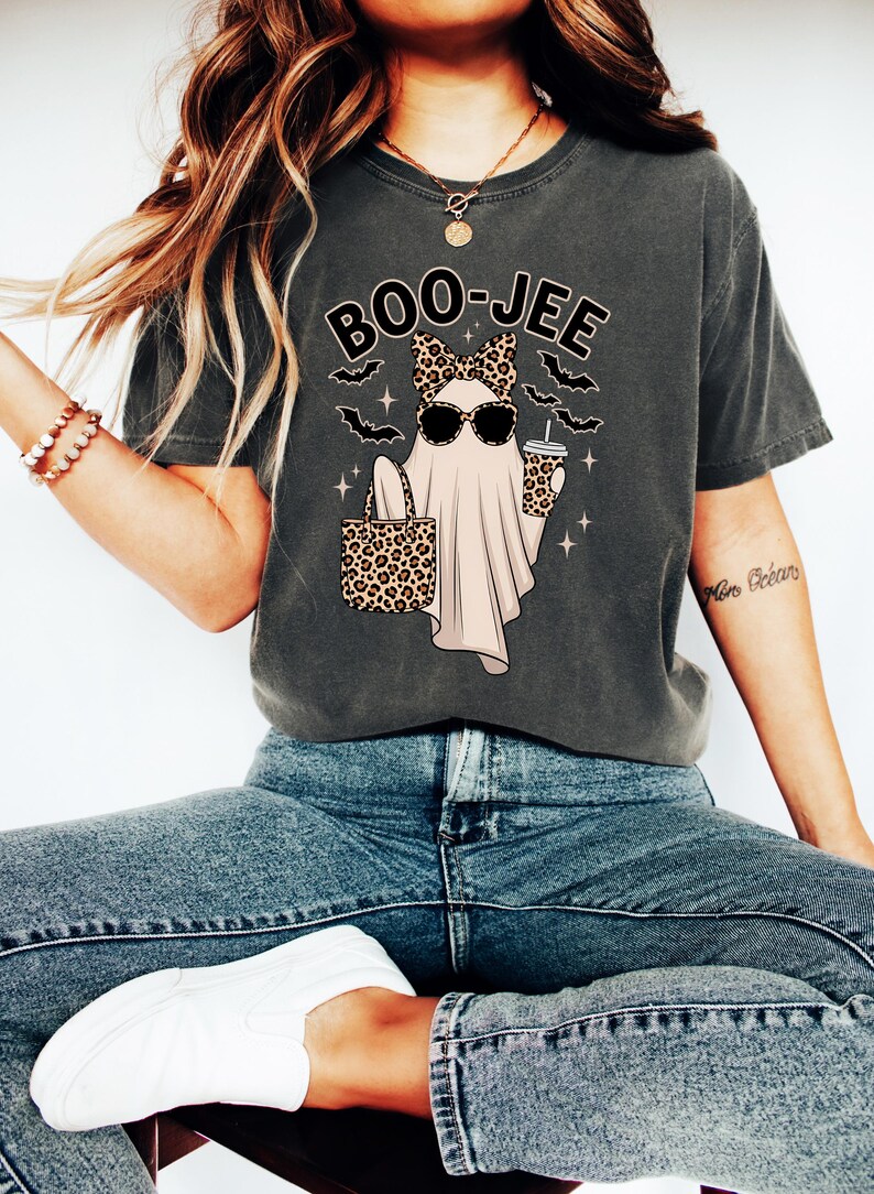 May include: Dark grey t-shirt with a Halloween graphic. The design shows a ghost wearing sunglasses and a leopard print bow, holding a leopard print bag and a coffee cup. The text "BOO-JEE" is above the ghost.