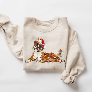 May include: A beige sweatshirt with a brown and white boxer dog wearing a red Santa hat and tangled in Christmas lights.