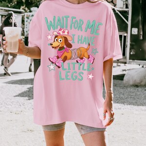 Puede incluir: Una camiseta rosa claro con el gr&aacute;fico de un perro salchicha marr&oacute;n con cuerpo de muelle, que lleva un sombrero de vaquero rosa, un pa&ntilde;uelo rosa y botas de vaquero rosas. El texto dice WAIT FOR ME I HAVE LITTLE LEGS, rodeado de estrellas rosas y azules.