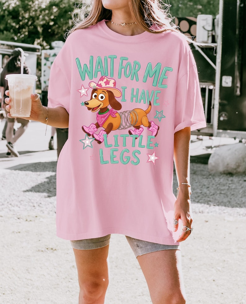 Puede incluir: Una camiseta rosa claro con el gr&aacute;fico de un perro salchicha marr&oacute;n con cuerpo de muelle, que lleva un sombrero de vaquero rosa, un pa&ntilde;uelo rosa y botas de vaquero rosas. El texto dice WAIT FOR ME I HAVE LITTLE LEGS, rodeado de estrellas rosas y azules.