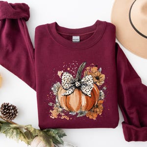 May include: Burgundy sweatshirt featuring a pumpkin graphic with a polka dot bow and floral accents. The pumpkin is orange with a green stem. A tan hat with a black band is in the background. Autumn-themed apparel.