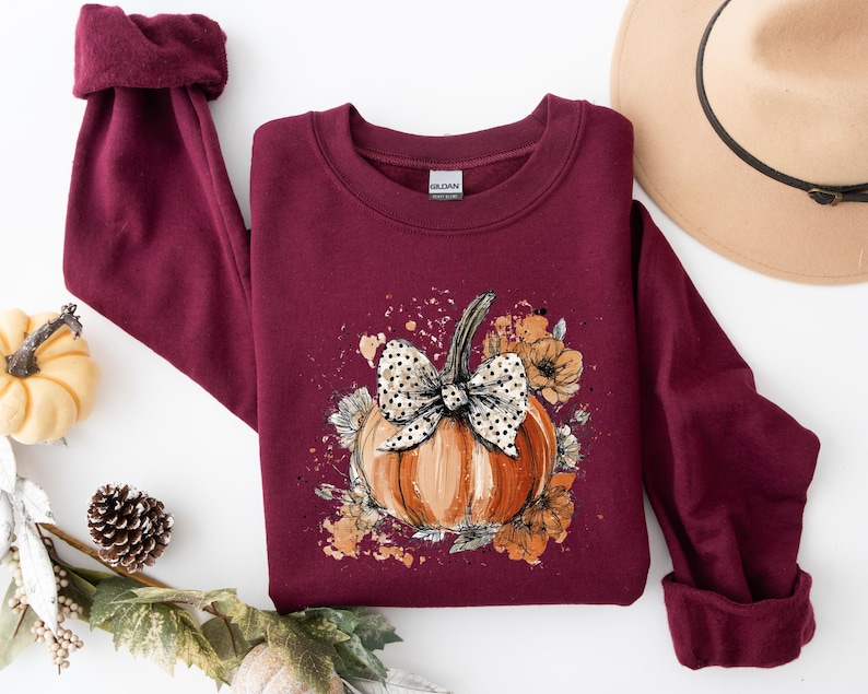 May include: Burgundy sweatshirt featuring a pumpkin graphic with a polka dot bow and floral accents. The pumpkin is orange with a green stem. A tan hat with a black band is in the background. Autumn-themed apparel.