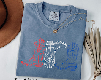 Peut inclure: Un t-shirt bleu jean avec un motif de trois bottes de cow-boy rouges, blanches et bleues. Le t-shirt a un col rond et des manches courtes. Un collier en or avec un pendentif est drapé sur le t-shirt. Le texte "Blue Jean" est en bas.