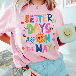 May include: A light pink t-shirt with the text "BETTER DAYS ARE ON THE WAY" in colorful letters and graphics. The shirt features a smiling sun, bees, and flowers. A white mug with a yellow smiley face is held in the hand.