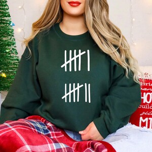 May include: A dark green sweatshirt with white tally marks printed on the front. The sweatshirt is part of a holiday theme, with a Christmas tree and festive pillows in the background. The person is wearing blue jeans and sitting on a red plaid blanket.