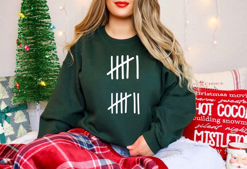May include: A dark green sweatshirt with white tally marks printed on the front. The sweatshirt is part of a holiday theme, with a Christmas tree and festive pillows in the background. The person is wearing blue jeans and sitting on a red plaid blanket.
