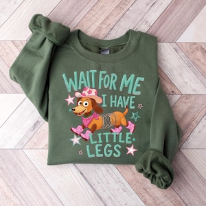 Puede incluir: Una sudadera de cuello redondo color verde oliva con un gr&aacute;fico de un perro salchicha marr&oacute;n con cuerpo de resorte met&aacute;lico, que lleva un sombrero de vaquero, pa&ntilde;uelo y botas rosas, con el texto "WAIT FOR ME I HAVE LITTLE LEGS" y estrellas.