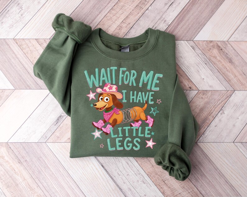 Puede incluir: Una sudadera de cuello redondo color verde oliva con un gr&aacute;fico de un perro salchicha marr&oacute;n con cuerpo de resorte met&aacute;lico, que lleva un sombrero de vaquero, pa&ntilde;uelo y botas rosas, con el texto "WAIT FOR ME I HAVE LITTLE LEGS" y estrellas.