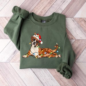 May include: A green Christmas sweatshirt with a brown and white boxer dog wearing a Santa hat and tangled in Christmas lights.