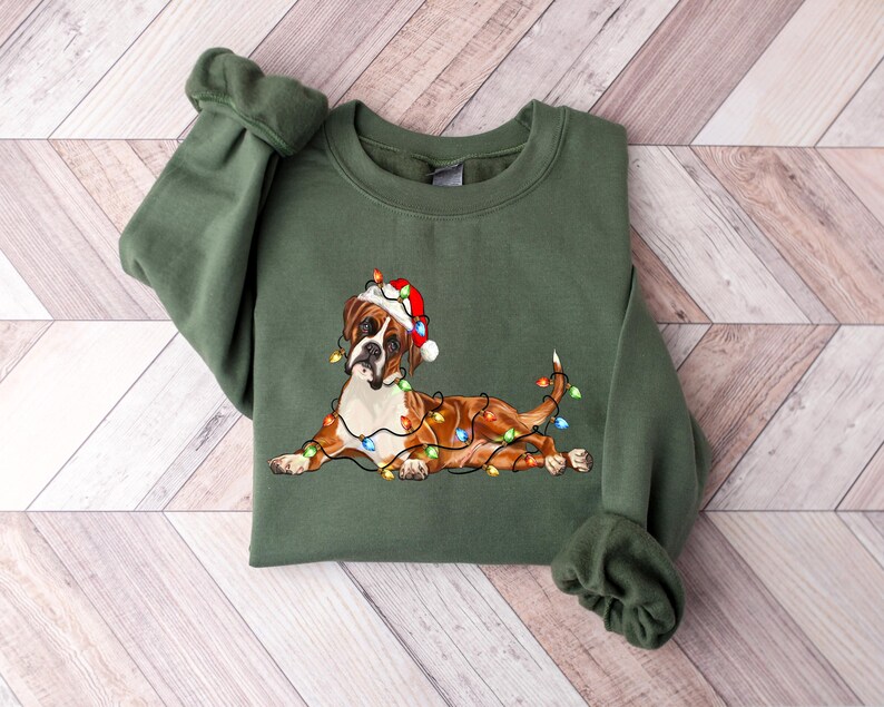 May include: A green Christmas sweatshirt with a brown and white boxer dog wearing a Santa hat and tangled in Christmas lights.
